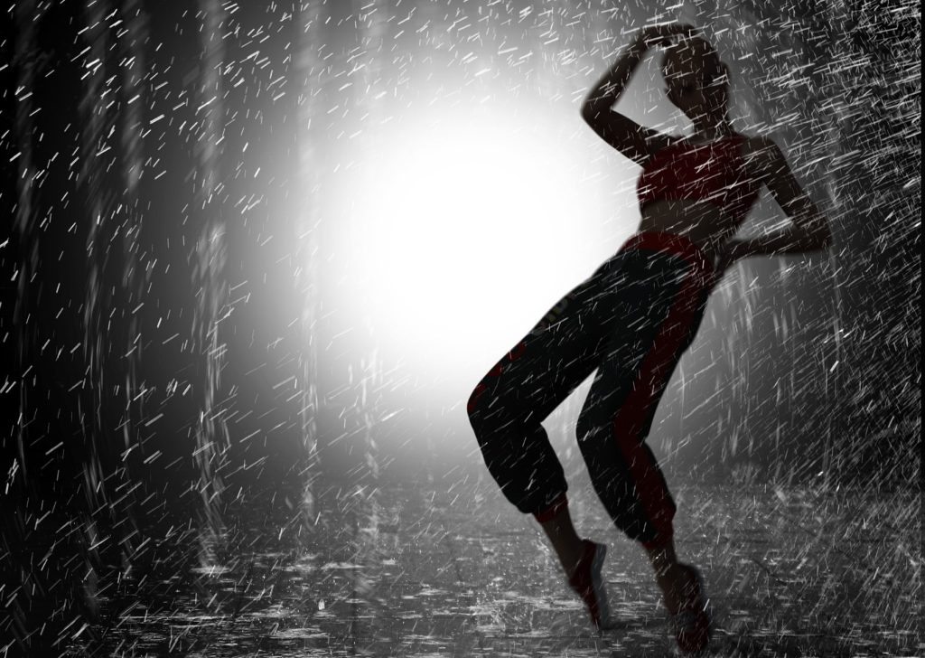 Silhuetten av en person som danser i regn med en sterk lyskaster bak seg.