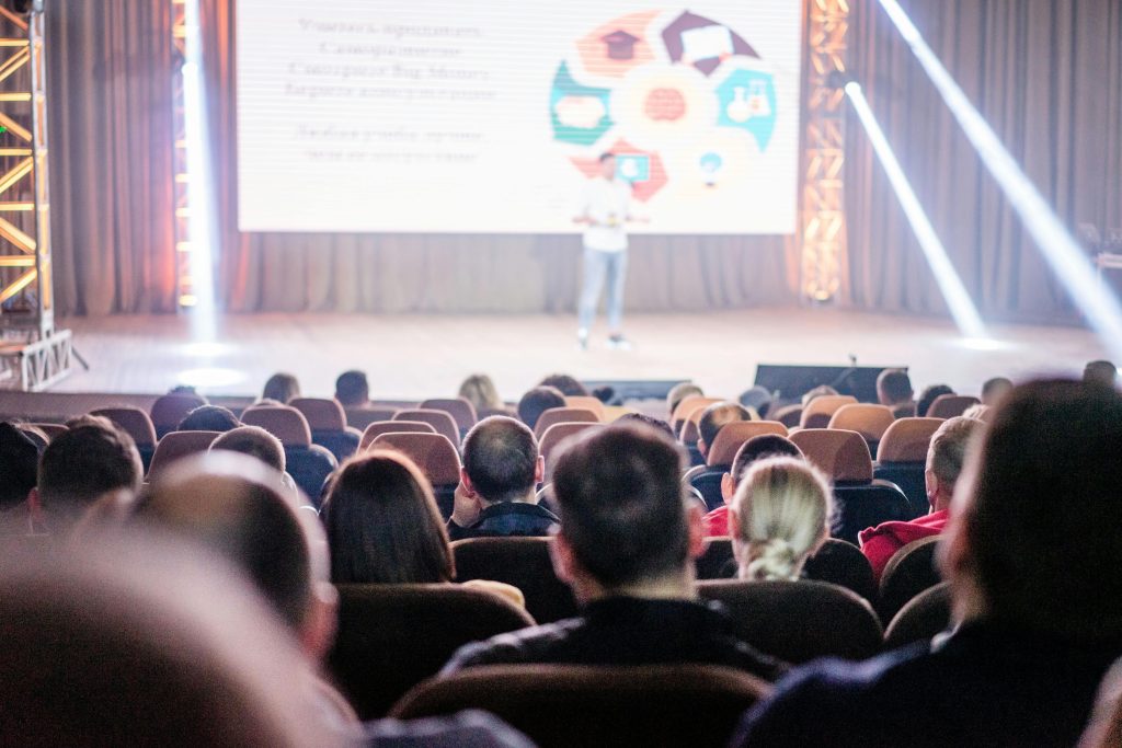 En foredragsholder på en scene med presentasjon på storskjerm, publikum sitter i salen.